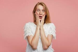 Amazed young pretty lady with foxy hair rounding her green-grey eyes while looking surprisedly to camera and keeping her face with raised hands, posing over pink background photo