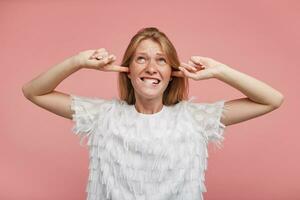 Displeased young attractive redhead lady with casual hairstyle keeping forefingers in her ears while trying to avoid annoying sounds, biting underlip while standing over pink background photo