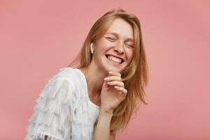 Good looking cheerful young redhead lady with natural makeup holding her chin with raised hand and laughing happily with closed eyes, isolated over pink background photo