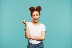 Flirty young attractive curly woman with bun hairstyle looking jocosely at camera and showing her tongue, wearing white basic t-shirt and jeans while posing over blue background photo