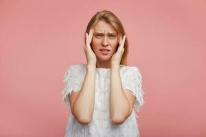 Upset young pretty female with foxy hair squinting her green-grey eyes and holding fingers on temples while suffering from strong headache, standing against pink background photo