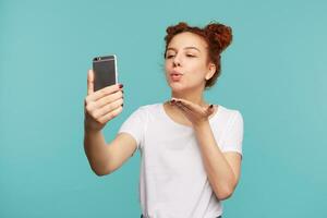 Portrait of young lovely brown haired curly lady with natural makeup folding her lips and blowing air kiss at camera while making photo of herself on her smartphone, isolated over blue background