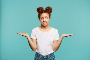 Bewildered young attractive redhead lady with casual hairstyle keeping her palms raised while looking confusedly aside, standing against blue background photo