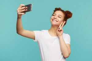 retrato de joven alegre Rizado morena hembra acuerdo teléfono inteligente en elevado mano mientras haciendo selfie y demostración felizmente paz gesto, aislado terminado azul antecedentes foto