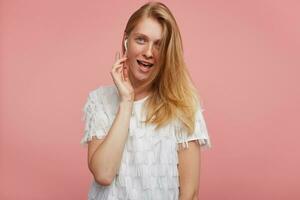 Excited young attractive redhead woman with casual hairstyle holding raised hand on earpiece and looking joyfully at camera with wide mouth opened, isolated over pink background photo