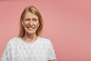 Confused young pretty redhead female frowning her face and showing teeth while grimacing her face, wearing white festive t-shirt while standing over pink background photo