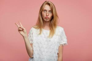 Funny shot of young lovely female with foxy hair raising fingers with peace sign and making faces while standing over pink background, dressed in white festive t-shirt photo
