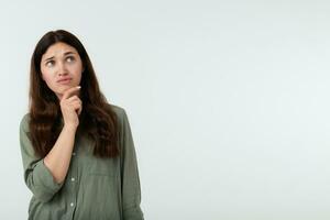 Pensive young pretty long haired brunette lady with casual hairstyle holding her chin with raised hand while looking thoughtfully upwards, standing over white background photo