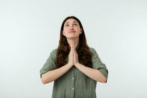 Desperate young brown haired lady with casual hairstyle frowning her eyebrows while looking worringly upwards, keeping palms together while posing over white background photo