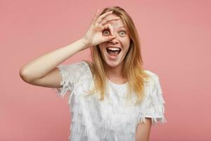 Joyful young beautiful woman with foxy hair raising ok gesture to her face and looking happily at camera and smiling happily with wide mouth opened, isolated over pink background photo