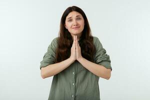 Indoor photo of pretty young long haired brunette lady looking hopefully at camera while folding raised palms in praying gesture, isolated over white background