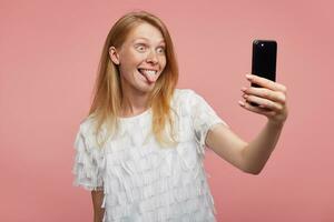 Funny shot of young joyful redhead lady sticking out her tongue and rounding green-grey eyes while making photo of herself with her mobile phone, isolated over pink background