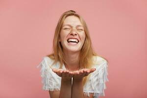 Studio photo of young joyful female with foxy hair raising her palms while standing over pink background, keeping eyes closed while smiling widely, being in high spirit