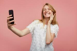 Indoor shot of joyful young lovely female with foxy hair showing her white perfect teeth while smiling widely, making selfie with her smartphone while standing over pink background photo