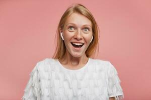 Portrait of young joyful lovely redhead woman with casual hairstyle wearing earphones while standing over pink background, rounding her green-grey eyes while looking happily upwards photo