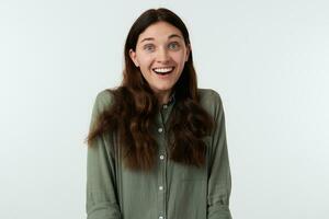 Overjoyed young lovely brown haired woman with natural makeup raising her eyebrows while looking cheerfully at camera with broad smile, isolated over white background photo