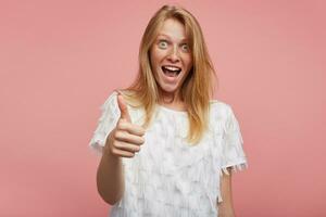 Overjoyed young lovely female with foxy hair showing hand with raised thumb and looking excitedly at camera with opened mouth, standing against pink background photo