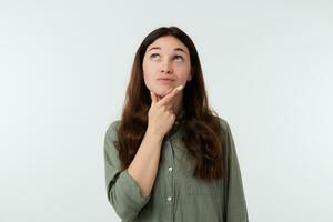 Puzzled young lovely long haired lady with natural makeup holding raised hand on her chin while looking pensively upwards, being concerned about something while posing over white background photo