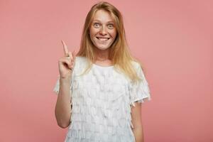 Overjoyed young pretty woman with foxy hair raising excitedly forefinger in having idea sign and looking surprisedly at camera with broad smile, isolated over pink background photo