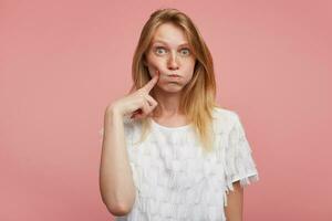 Indoor photot of young attractive redhead lady puffing out her cheeks and keeping foreginger on her face while looking excitedly to camera, standing against pink background photo