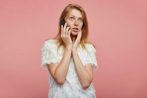 Studio photo of open-eyed pretty young redhead lady holding palm on her face while posing over pink background, looking surprisedly ahead while having phone conversation