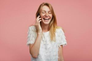 Good looking young lovely redhead lady with natural makeup keeping her eyes closed while laughing happily, having phone talk while standing over pink background photo