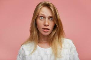 de cerca de confuso joven pelirrojo dama con casual peinado mirando a cámara con amplio ojos abrió mientras en pie terminado rosado antecedentes en blanco festivo camiseta foto