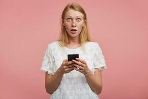 Shocked young attractive female with foxy hair rounding her green-grey eyes while looking amazedly at camera, holding mobile phon in raised hands while posing over pink background photo