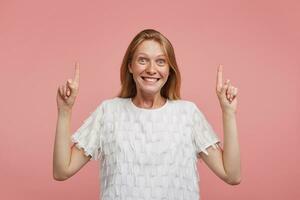 Excited young beautiful joyful female with foxy hair looking at camera with agitated face and smiling broadly, standing over pink background with raised index fingers photo