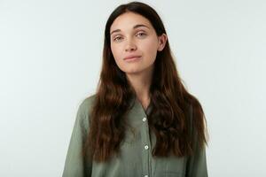 Pleasent looking young brown haired woman with natural makeup keeping her lips folded while standing against white background in green casual shirt photo