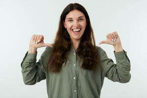 Indoor shot of young complacent long haired brunette lady laughing happily while showing on herself with raised hands, isolated against white background photo