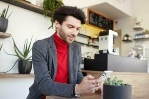 Side view of young handsome unshaved brunette male keeping mobile phone in his hands and looking positively on screen while waiting for his order in cafe photo