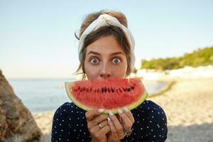 Young dark haired pretty woman in headband making fun over seaside view with slice of watermelon in her hands, looking at camera with wide eyes opened and raising eyebrows photo