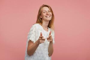 Indoor shot of young happy redhead woman with natural makeup pointing to camera with raised forefingers and smiling cheerfully while posing over pink background photo