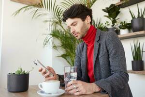 Indoor shot of young handsome bearded brunette man with trendy haircut reading message on his mobile phone while having cup of coffee in city cafe during lunch time photo