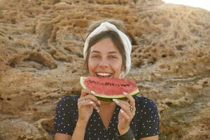 Happy beautiful young dark haired female in headband with casual hairstyle posing over big yellow stone on sunny warm day, eating watermelon and looking cheerfully to camera photo