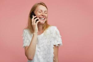 retrato de bueno mirando joven contento mujer con astuto pelo acuerdo teléfono inteligente en elevado mano y sonriente alegremente mientras teniendo bonito conversación, aislado terminado rosado antecedentes foto