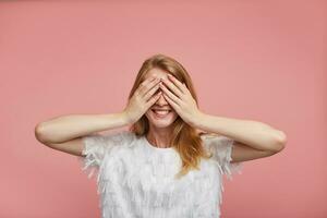 Studio photo of happy lovely young redhead lady in white elegant blouse keeping raised palms on her face and smiling cheerfully while posing over pink background