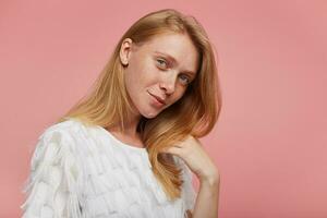 Portrait of beautiful young redhead female with casual hairstyle wearing white festive blouse while standing over pink background, keeping her lips folded while looking gently at camera photo