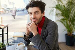 Portrait of beautiful young bearded brunette man looking positively at camera with charming smile and leaning his head on raised hands while sitting in city cafe photo