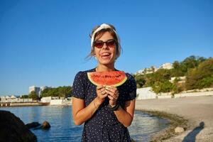 Funny outdoor shot of charming young dark haired woman with casual hairstlyle standing over sea bay, keeping piece of watermelon in hands and showing tongue happily photo