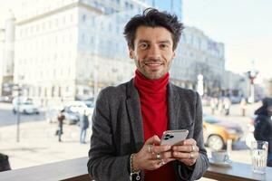 retrato de positivo joven atractivo marrón peludo masculino con de moda Corte de pelo sonriente alegremente a cámara y acuerdo teléfono inteligente en elevado manos, esperando para amigos después trabajo en verano terraza foto