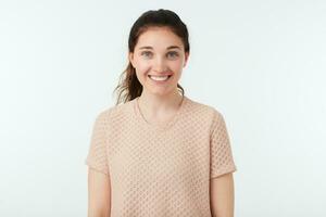 Cheerful young beautiful brunette female with casual hairstyle looking gladly at camera with charming smile while posing over white background in nude t-shirt photo