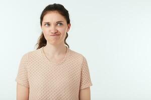 Horizontal shot of young green-eyed pretty brunette female grimacing her face while looking confusedly aside, standing over white background with hands down photo