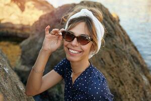 hermosa joven morena dama en de moda Gafas de sol y blanco venda posando al aire libre en soleado día, sonriente alegremente a cámara mientras sentado en grande Roca foto