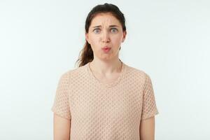 Discouraged young pretty brown haired female frowning her eyebrows while looking confusedly at camera, dressed in beige t-shirt while posing over white background photo