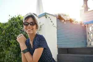 positivo encantador joven hembra en Clásico de moda Gafas de sol y blanco venda posando al aire libre en soleado día, sentado terminado escalera en patio interior, plegable elevado manos y sonriente alegremente foto