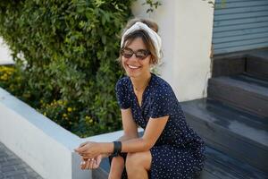 hermosa joven hembra con marrón pelo vistiendo Clásico de moda Gafas de sol, sentado en escalera terminado patio interior, mantener manos en su rodillas y mirando felizmente a cámara foto