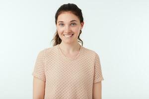 Studio photo of young charming dark haired lady with ponytail hairstyle showing her perfect white teeth while smiling happily, standing over white background