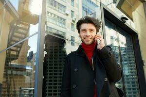 al aire libre foto de joven bonito sin afeitar morena hombre en de moda ropa haciendo llamada con su teléfono inteligente y mirando afirmativamente a cámara con agradable sonrisa